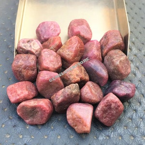 May include: A collection of rough, pink ruby gemstones in various sizes and shapes. The gemstones are displayed in a metal tray.