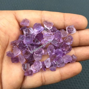May include: A pile of small, rough-cut amethyst crystals in various shades of purple. The crystals are scattered on a hand, showcasing their natural beauty.