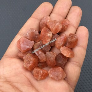 K&ouml;nnte beinhalten: Ein Haufen kleiner, rauer, orange-roter Sunstone-Kristalle. Die Kristalle sind durchscheinend und haben einen leichten Glanz.