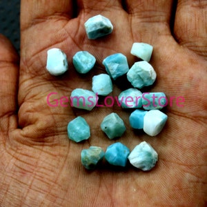May include: A collection of small, rough-cut gemstones in shades of light blue and white. The stones are held in a hand, showcasing their irregular shapes and natural textures. The overall aesthetic is organic and earthy.