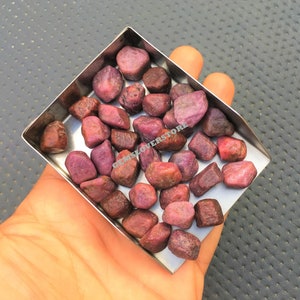 May include: A collection of small, rough, pink ruby gemstones in a metal tray. The gemstones are various shades of pink and have a natural, unpolished look.