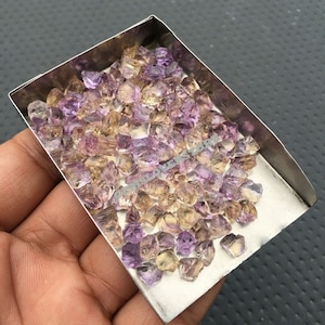 May include: A small metal tray filled with a variety of small, faceted, amethyst and citrine gemstones. The gemstones are a mix of purple, yellow, and orange.