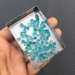May include: A small metal tray filled with many small, faceted, teal blue gemstones. The tray is being held in a person's hand.