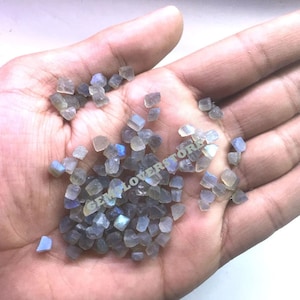 May include: A close-up of a hand holding a pile of small, iridescent, gray labradorite gemstone chips.