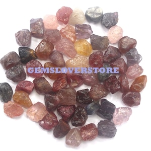 Op de afbeelding: Een verzameling losse, ruw geslepen edelstenen in verschillende tinten rood, roze, oranje en blauw. De edelstenen zijn allemaal ongeveer even groot en van dezelfde vorm, en ze zijn op een witte achtergrond in een stapel gerangschikt. De tekst "GEMSLOVERSTORE" is zichtbaar in de afbeelding.
