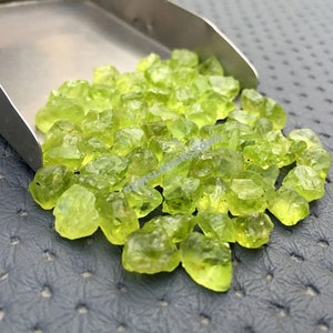 Puede incluir: Un montón de pequeñas piedras preciosas de peridoto verde y rugosas. Las piedras preciosas están esparcidas sobre una superficie gris texturizada.