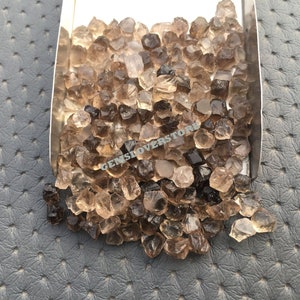 May include: A close-up of a pile of small, brown, faceted quartz crystals. The crystals are in a metal scoop, and the background is a gray, textured surface.