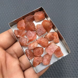 K&ouml;nnte beinhalten: Eine Sammlung von rohen, orange-roten Sunstone-Kristallen in einer Metallschale. Die Kristalle sind in verschiedenen Gr&ouml;&szlig;en und Formen, einige haben einen leichten Schimmer.