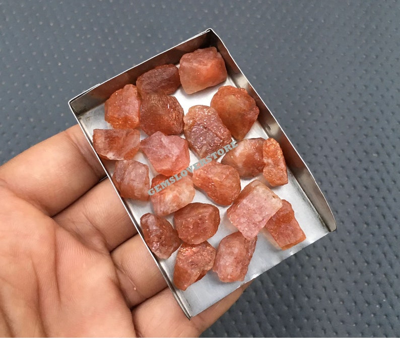 K&ouml;nnte beinhalten: Eine Sammlung von rohen, orange-roten Sunstone-Kristallen in einer Metallschale. Die Kristalle sind in verschiedenen Gr&ouml;&szlig;en und Formen, einige haben einen leichten Schimmer.