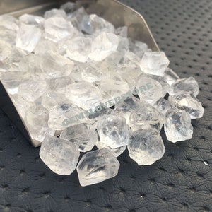 Pode incluir: Uma pá cheia de cristais de quartzo transparentes e facetados. Os cristais são de vários tamanhos e formas, e estão a derramar-se da pá para uma superfície preta.