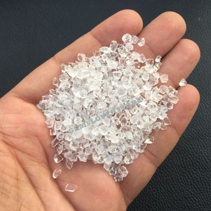 Puede incluir: Una mano sosteniendo un montón de cristales de vidrio transparentes y facetados. Los cristales son pequeños y de tamaño uniforme, y brillan a la luz.