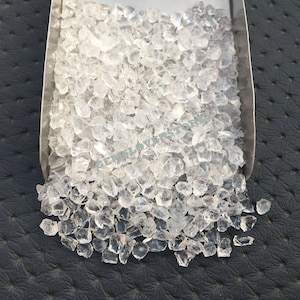 Puede incluir: Una imagen de primer plano de un montón de cristales de cuarzo transparentes. Los cristales son pequeños y tienen una variedad de formas y tamaños. Se están derramando de una pala de metal sobre una superficie negra.