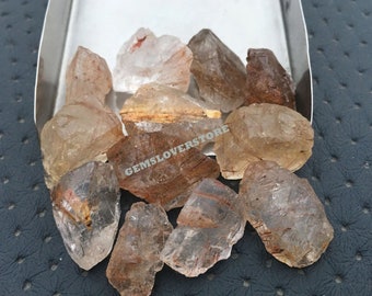 May include: A collection of raw, translucent quartz crystals with reddish-brown inclusions. The crystals are various sizes and shapes, and some have a slight golden hue.
