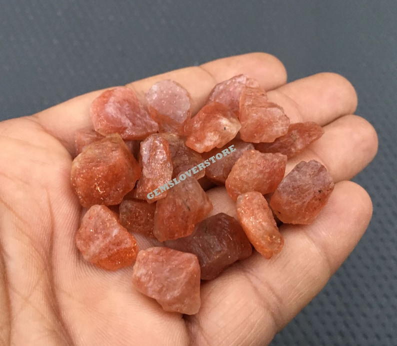 K&ouml;nnte beinhalten: Eine Sammlung kleiner, rauer, orange-roter Sunstone-Kristalle. Die Kristalle sind durchscheinend und haben ein glitzerndes, funkelndes Aussehen.
