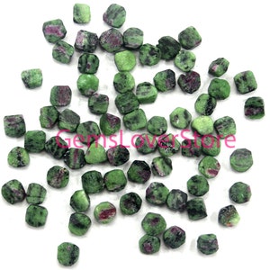 May include: A collection of green and pink gemstone beads, each with a faceted, octagonal shape. The beads are scattered on a white surface.