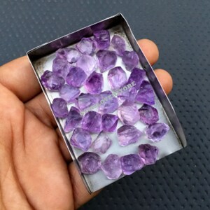 May include: A collection of raw amethyst crystals in various sizes and shapes, displayed in a small metal tray. The crystals are a deep purple colour and have a natural, rough texture.