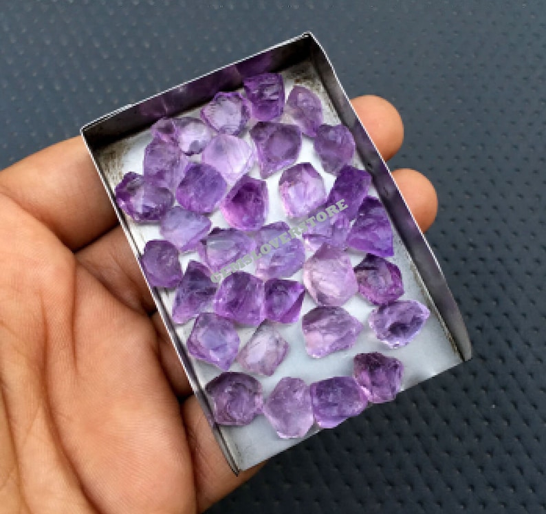 May include: A collection of raw amethyst crystals in various sizes and shapes, displayed in a small metal tray. The crystals are a deep purple colour and have a natural, rough texture.