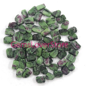 May include: A collection of tumbled ruby in zoisite stones. The stones are a mix of green and dark pink, with a rough, natural texture. The gemstones are scattered on a white surface.