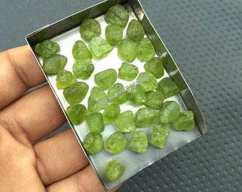 May include: A small metal tray filled with approximately 30 green peridot gemstones. The tray is being held in a person's hand.