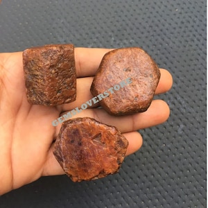 May include: Three rough, reddish-brown gemstones with a hexagonal shape. The gemstones are raw and have a natural, unpolished finish.