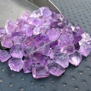 May include: A pile of small, rough-cut amethyst crystals in various shades of purple. The crystals are loose and scattered on a gray surface.