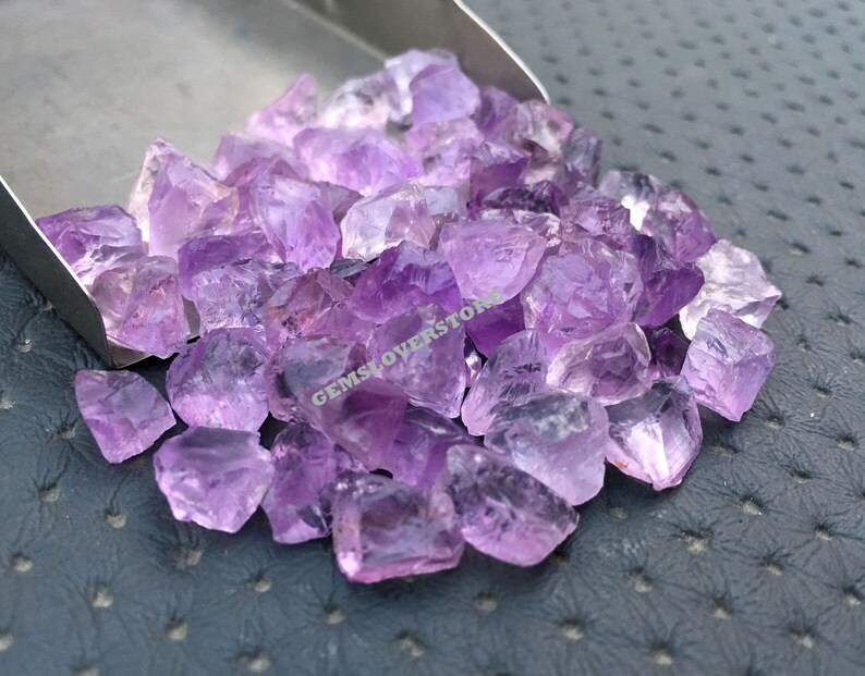 May include: A pile of small, rough-cut amethyst crystals in various shades of purple. The crystals are loose and scattered on a gray surface.