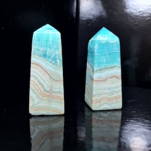 May include: Two blue-green gemstone obelisks with a layered pattern of tan, white, and brown. The obelisks have a rectangular base and a pointed top. The background is black.
