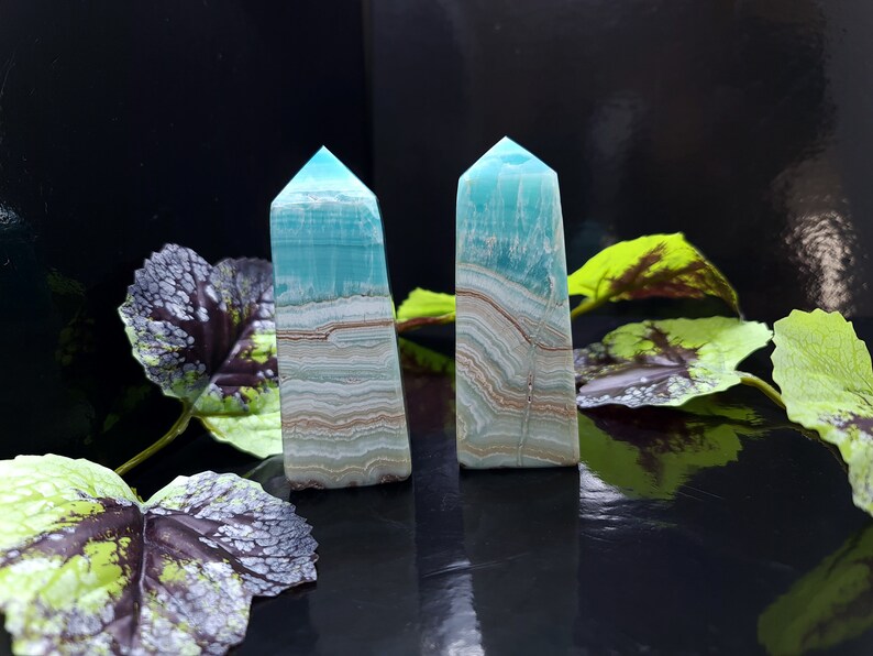 May include: Two turquoise and white banded crystal obelisks with a polished finish. The obelisks have a rectangular base and a pointed top. The background is black with green and purple leaves.