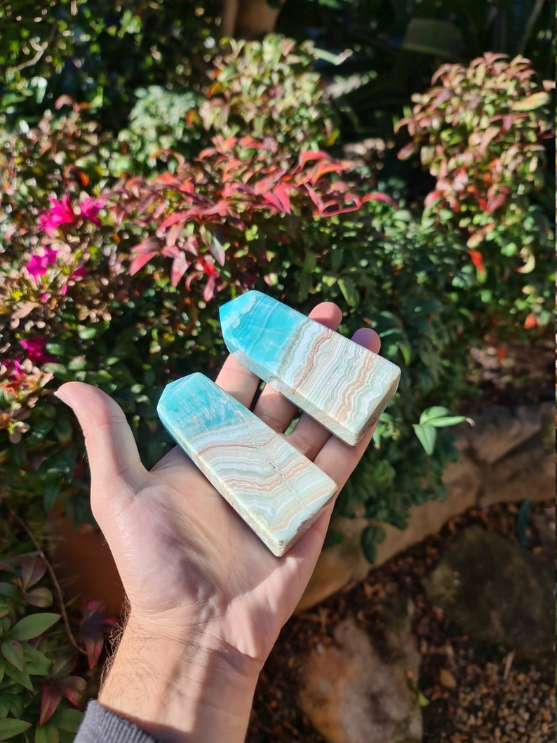 May include: Two rectangular, polished gemstone obelisks. The obelisks are light blue at the top and transition to a cream and tan striped pattern. The obelisks are held in a hand against a backdrop of green and red foliage.