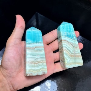 May include: Two blue and tan crystal obelisks. The obelisks have a rectangular shape with a pointed top. The blue color is at the top, transitioning to tan and white bands. The crystals are held in a hand.