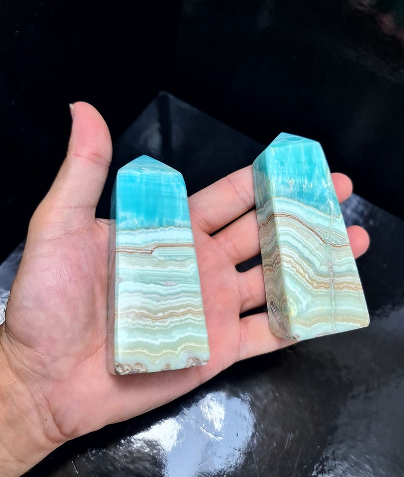 May include: Two blue and tan crystal obelisks. The obelisks have a rectangular shape with a pointed top. The blue color is at the top, transitioning to tan and white bands. The crystals are held in a hand.