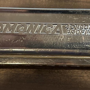 May include: A silver chromatic harmonica with the text "The 64 Chromonica" and "4 Chromatic Octaves Professional Model". The harmonica has a silver body with gold-colored end plates and a small silver mouthpiece.
