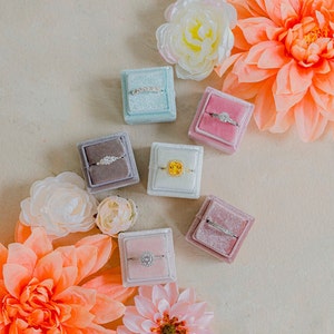 May include: Seven velvet ring boxes in various colors, including pink, blue, and gray, each containing a different ring. The boxes are arranged on a light beige surface with flowers.
