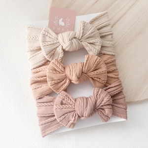 May include: Three knotted headbands in beige and pink hues are displayed. The headbands have a textured, ribbed design. A small pink card with a rabbit logo is visible. The headbands are presented on a white surface.