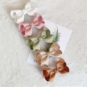 May include: Four fabric hair bows in different colours: white, pink, green, and brown. The bows are arranged in a row on a white surface.