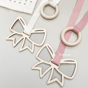 May include: Two wooden bow tie shapes with a ring attached to each. One bow tie is attached to a white ribbon and the other is attached to a pink ribbon.