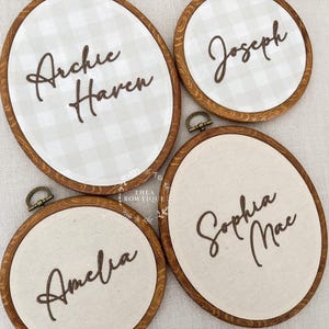 May include: Four wooden embroidery hoops containing fabric with names in dark brown script. Two hoops feature a light gray and white gingham pattern, while two feature solid cream fabric. The names Archie Haven, Joseph, Amelia, and Sophia Mae are displayed.
