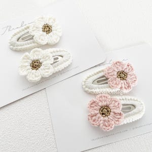 May include: Two pairs of handmade crochet flower hair clips. One pair features white flowers with gold centres, the other has pink flowers with gold centres. The clips are on white cards.