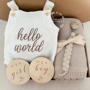May include: A baby gift set in a box, including a white knitted romper with "hello world" text, a beige blanket, a pacifier clip, and two wooden discs reading "it's a girl" and "it's a boy".