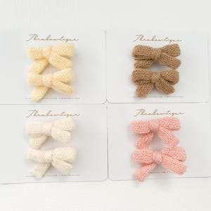 May include: Four sets of knitted hair bows, each set containing two bows. The bows are in four colours: yellow, brown, white, and pink.