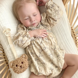 Könnte beinhalten: Ein Baby in einem cremefarbenen, blumigen Smocks mit langen Ärmeln, das in einem Weidenkörbchen liegt. Ein plüschiger brauner Teddybär, der an einem Schnullerclip befestigt ist, liegt auf der Bettwäsche.