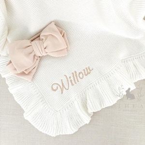May include: A white knitted baby blanket with a ruffled edge and the name "Willow" embroidered in gold thread. A blush pink bow is placed on top of the blanket.
