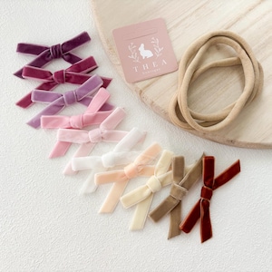 May include: An assortment of velvet hair bows in various colours, including purple, pink, and brown. The bows are arranged in a semi-circle on a white surface. A nude-coloured elastic headband is also included.