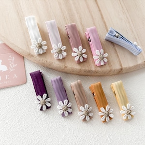 May include: A collection of colorful hair clips with daisy accents. The clips are in various colors, including white, pink, purple, and orange. A pink tag with a bunny logo and the text "THEA boutique" is also visible.