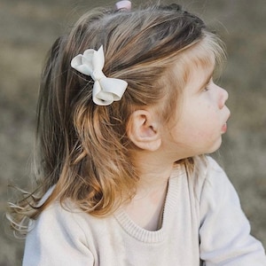 May include: A young girl with long, wavy blonde hair wearing a white sweater. She has a white bow in her hair and is looking to the right.