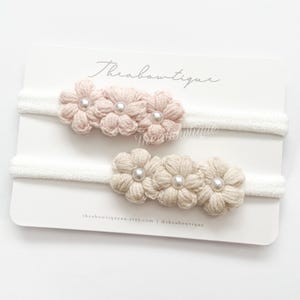 May include: Two sets of crocheted flower headbands. One set is pink and the other is beige. Each set has three flowers with a pearl in the centre. The headbands are white.