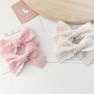 以下が含まれることがあります： ソフトピンクとクリーム色のレースのヘアボウ2セット。各リボンは、繊細なカットアウトパターンで複雑に設計されています。リボンは、「Theabout」というテキストとロゴが入った白いカードに表示されています。