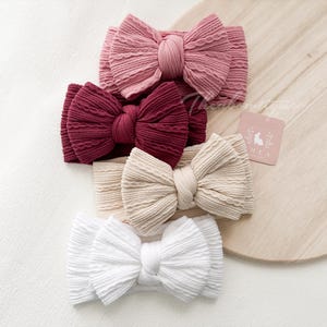 Könnte beinhalten: Vier strukturierte Baby-Stirnbänder in Rosa, Burgunderrot, Beige und Weiß. Jedes Stirnband hat eine große Schleife. Ein kleines Etikett mit einem Hasenlogo ist sichtbar.