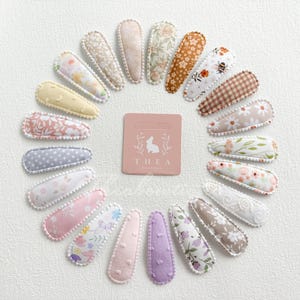 May include: A set of 16 floral patterned hair clips with a white trim. The clips are arranged in a circle on a white background. The text "THEA Boutique" is printed on a small card in the center of the circle.