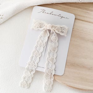 May include: A cream-coloured lace bow hair clip with long, flowing tails. The bow is attached to a white card with the text "Theabouitique" in cursive. The lace features a delicate floral pattern.